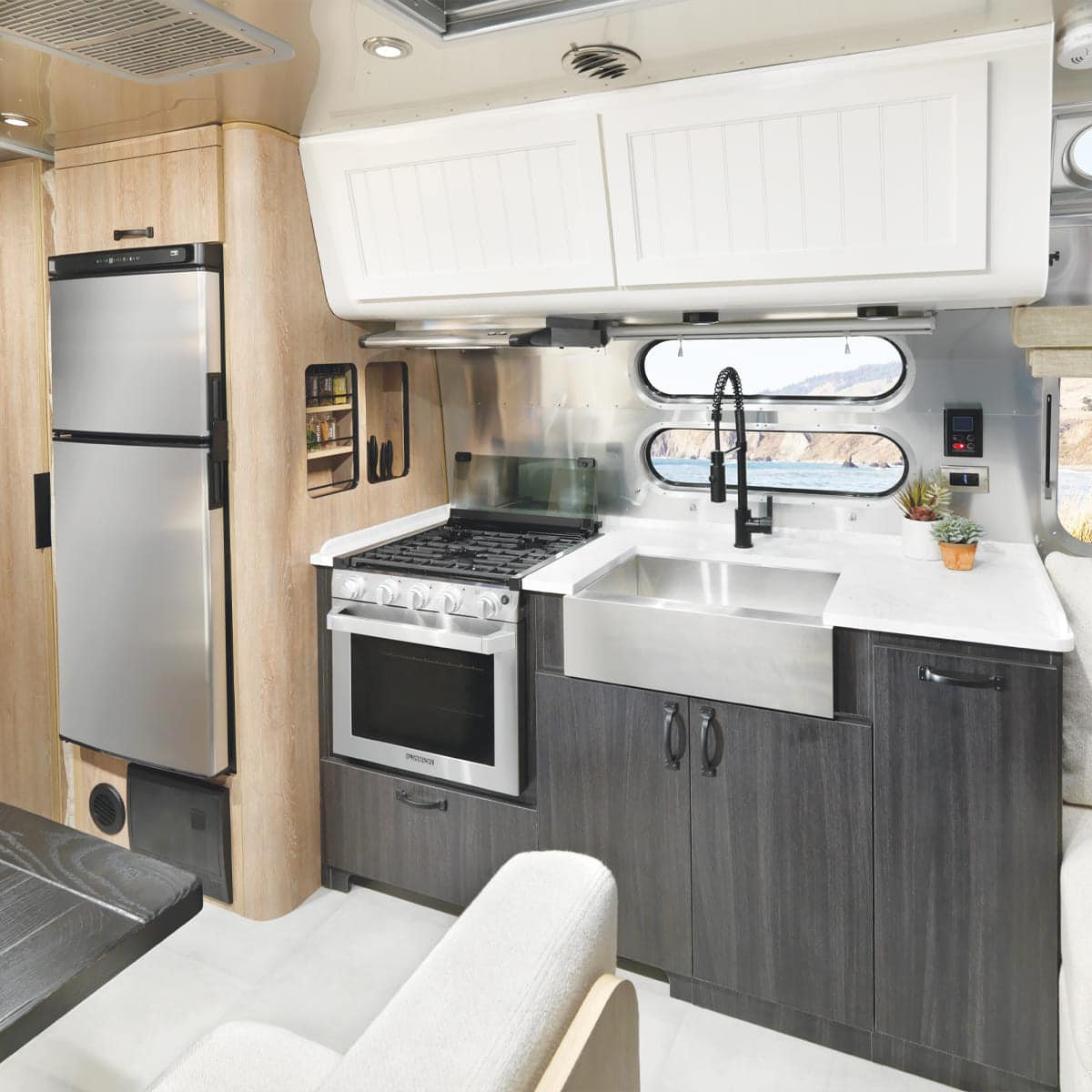 A modern Airstream RV kitchen features a stainless steel fridge, oven, and deep sink, with white and dark cabinets, a window above the sink, and a small plant on the countertop for a touch of Pottery-Barn style charm.