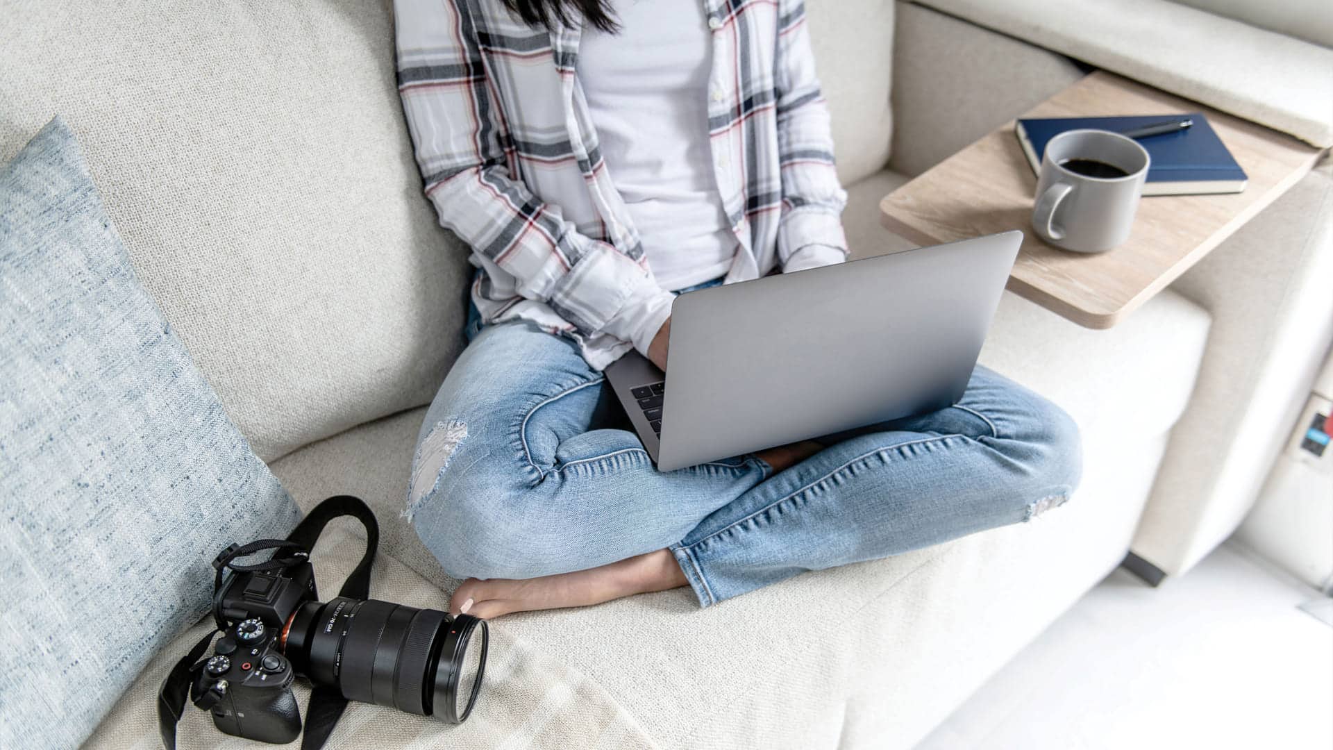 A person sits cross-legged on a Pottery-Barn couch using a laptop. A camera rests beside them, and a tray with coffee, notebook, and pen is on the armrest. The person wears jeans and a plaid shirt.