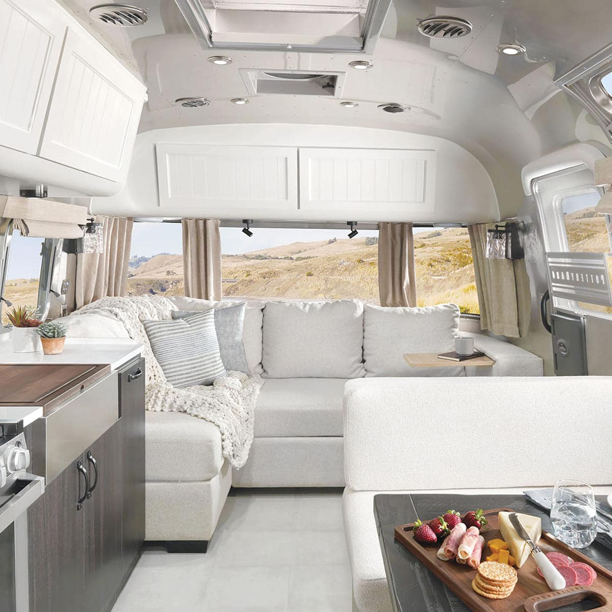 Interior of a modern Airstream RV with a light-colored L-shaped Pottery Barn sofa, cozy blankets, large windows revealing scenic views, kitchen area to the left, and a tray with snacks and drinks on a small table in the foreground.