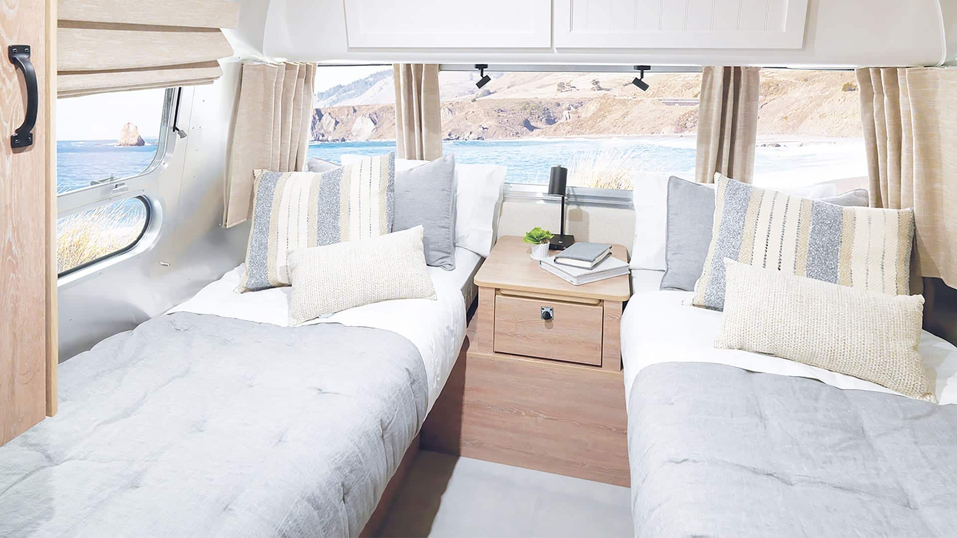 Two twin beds with gray Pottery-Barn bedding and striped pillows inside a bright Airstream camper, separated by a wooden nightstand. Large windows reveal a scenic coastal view with ocean waves and rocky cliffs outside.