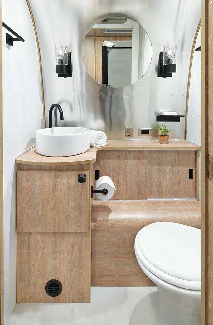 Modern, compact bathroom with wood accents, Pottery-Barn round mirror, vessel sink, black faucet, wall lights, small potted plant, and white toilet—all neatly arranged in a stylish Airstream-inspired space.