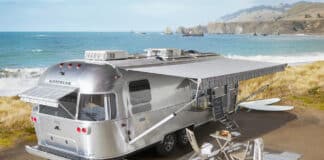 A silver Pottery-Barn Airstream trailer with an extended awning is parked by the ocean. Surfboards, outdoor chairs, and a small table set on a striped rug create a cozy setup overlooking the beach and cliffs in the background.