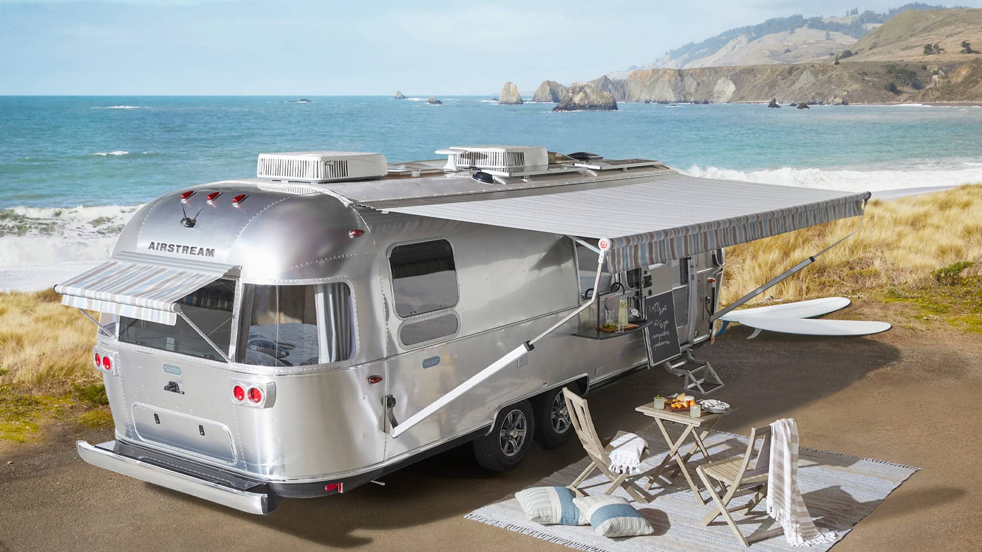 A silver Pottery-Barn Airstream trailer with an extended awning is parked by the ocean. Surfboards, outdoor chairs, and a small table set on a striped rug create a cozy setup overlooking the beach and cliffs in the background.