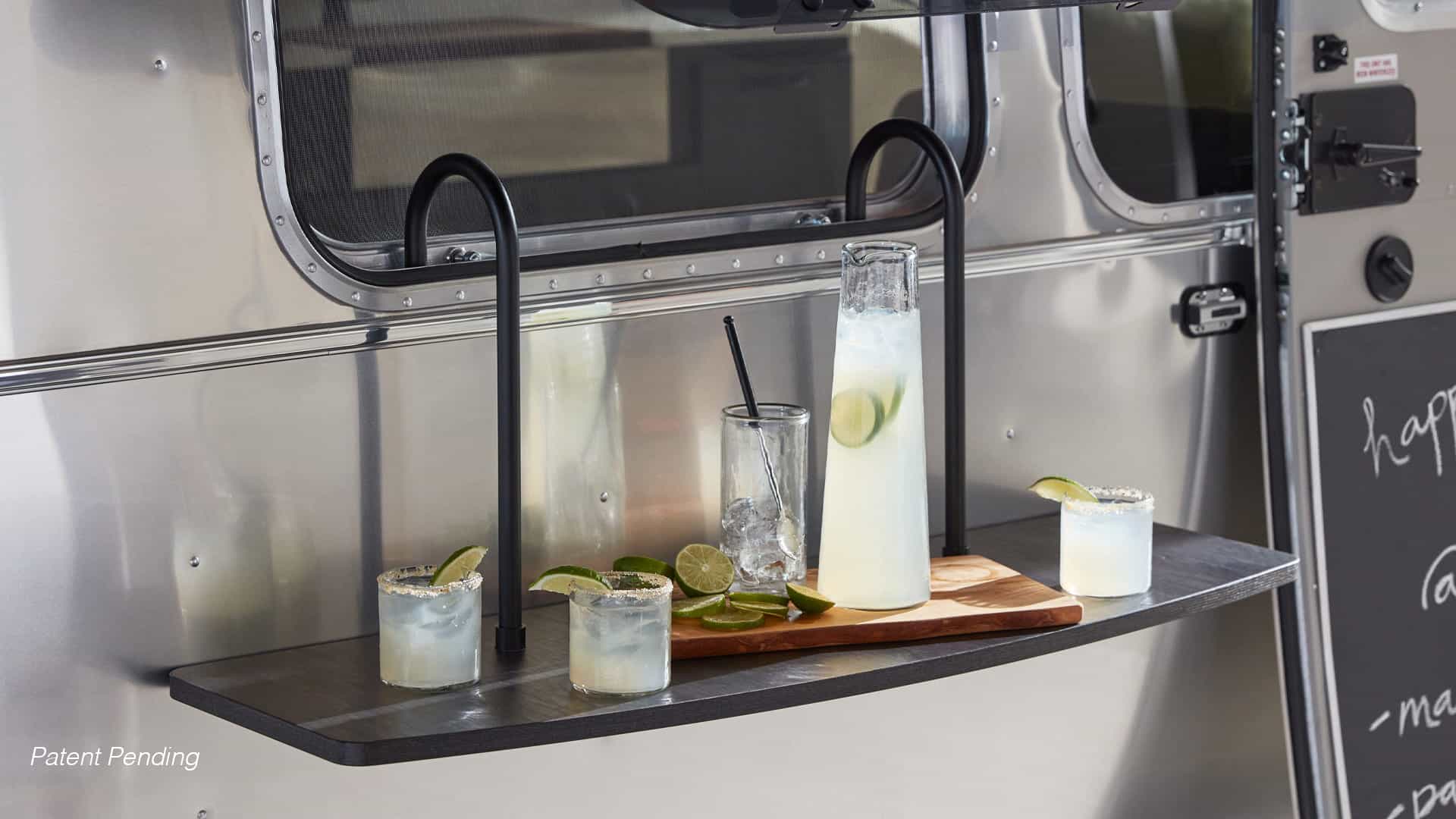 A small outdoor shelf attached to an Airstream’s metallic surface holds two cocktails, a tall glass pitcher of lemonade, a glass with ice and straw, sliced limes, and a cutting board—perfect for Pottery-Barn-inspired entertaining.