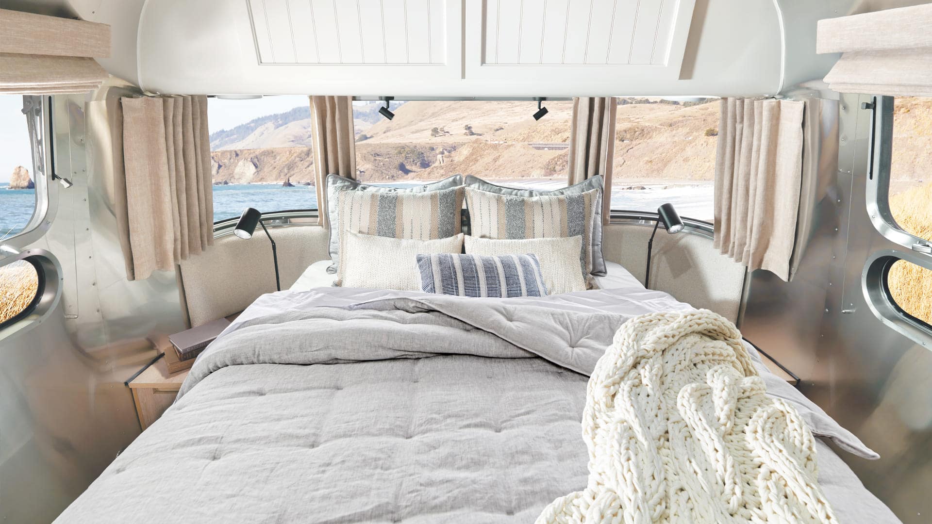 A cozy bed with Pottery Barn neutral bedding and pillows is inside an Airstream trailer, with windows showing a scenic ocean and hillside view. Beige curtains frame the windows, and a chunky knit blanket lies on the bed.