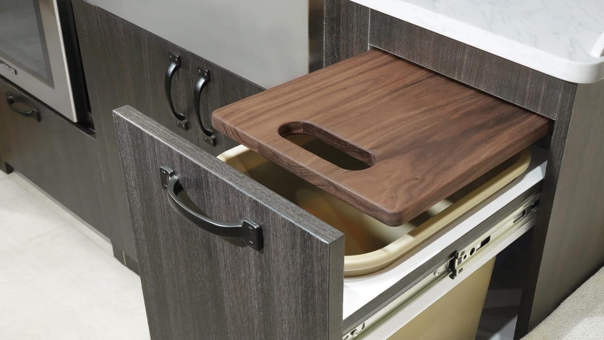 A Pottery-Barn inspired kitchen with a pull-out trash bin beneath a sliding wooden cutting board, allowing food scraps to be easily disposed of while preparing meals.