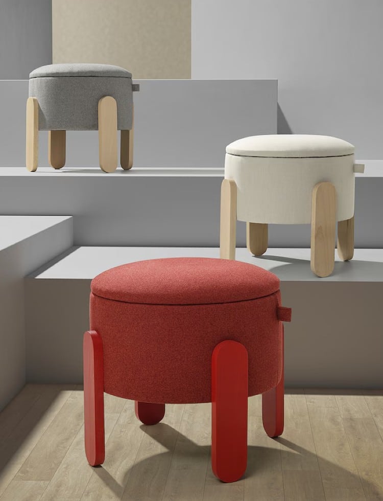 Three round, modern ottomans with wooden legs in gray, white, and red are arranged on light gray steps and a wooden floor in a minimalist setting.