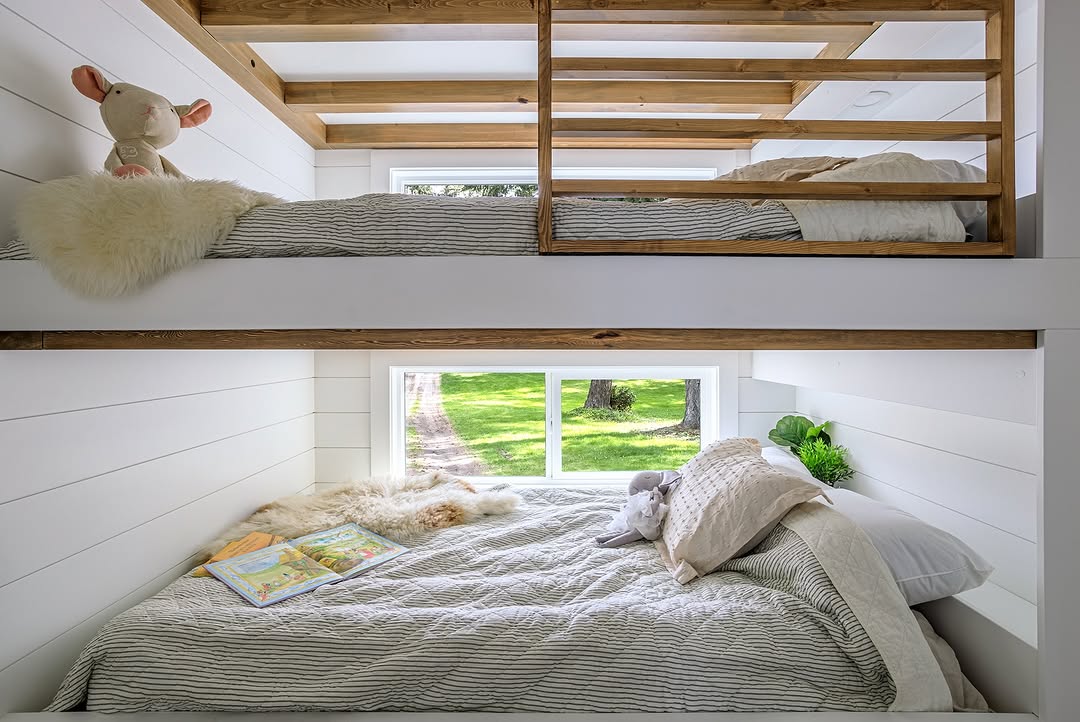 A cozy bunk bed with wood accents in a bright room. The lower bunk has pillows, a book, and fur throw, while the upper bunk has a stuffed animal and a furry cushion. Large windows let in natural light and show a green yard outside.