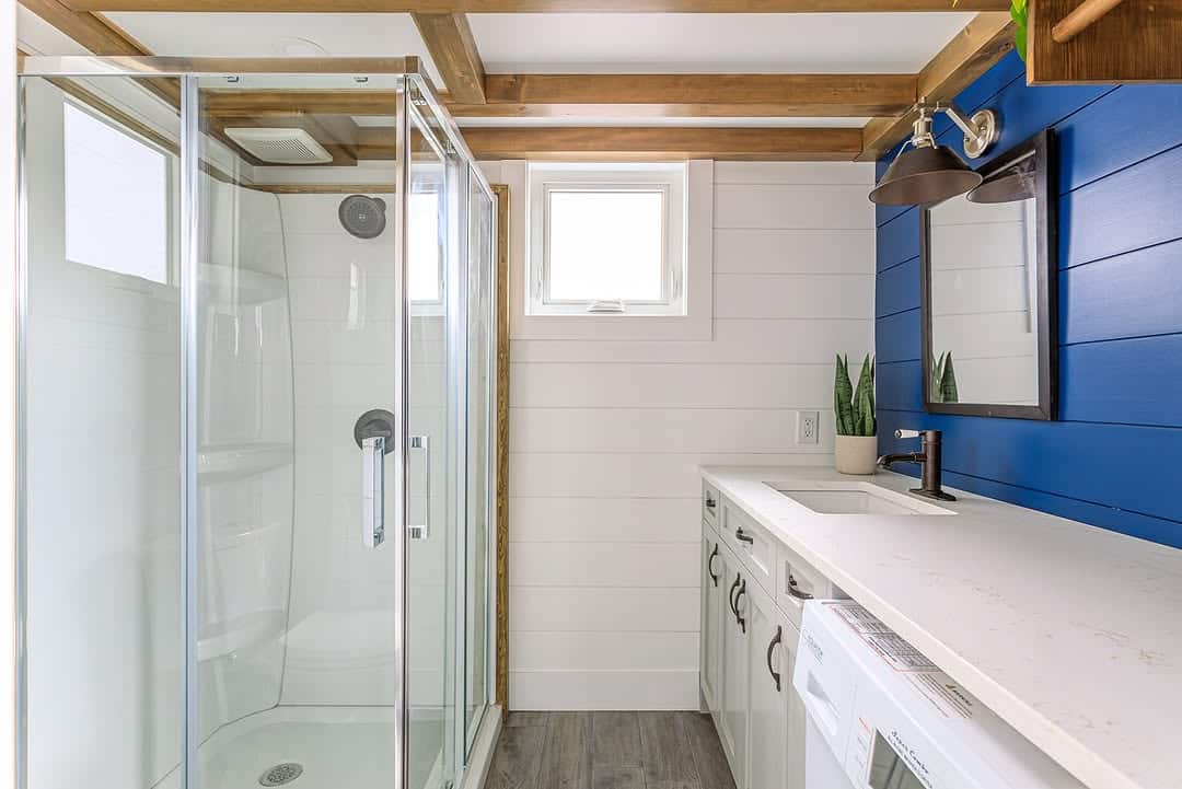 Modern bathroom with a glass shower, white vanity with sink, green plant, blue accent wall with mirror and light, and a washing machine under the counter, featuring wood ceiling beams and a small window.