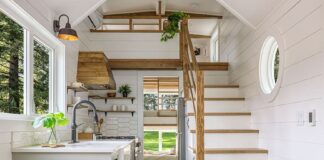 Bright, modern tiny house interior with white walls, wood accents, kitchen, staircase to loft, round window, plants, a small dining area with a chair, and a cozy living space with a gray sofa. Sunlight streams in through windows.