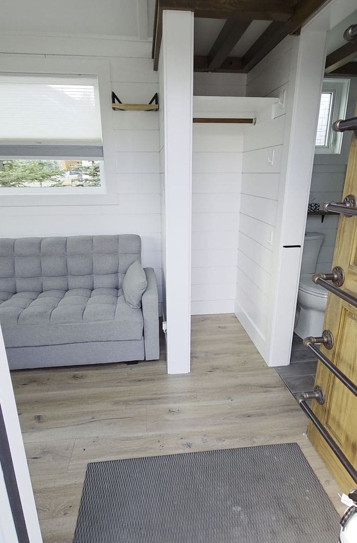 A small, modern room with light wood floors features a gray upholstered sofa on the left, a narrow open closet in the center, and a partial view of a bathroom with a toilet on the right.
