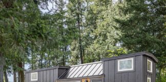 A modern tiny house on wheels with dark wood siding, white-trim windows, and a small wooden porch sits on a grassy area among tall trees, with potted plants and two chairs outside.