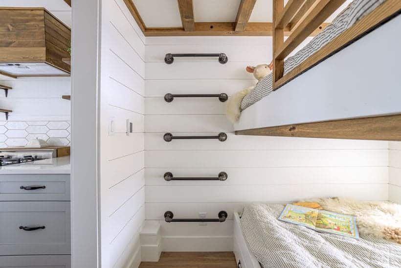 A cozy room features a built-in bunk bed with a stuffed animal on the top bunk, metal rungs forming a ladder on a white shiplap wall, and a book lying open on the lower bed; a kitchen is visible to the left.