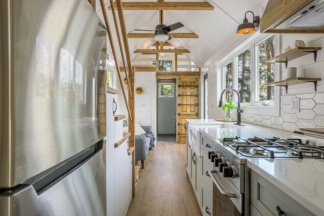 A modern tiny house kitchen with white cabinets, stainless steel appliances, a gas stove, open wooden shelves, and a loft above. Large windows let in natural light, and a wooden door is at the end of the space.