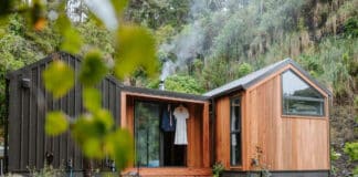 A modern tiny house with wood and black siding sits among rocks and greenery. Smoke rises from a chimney, and clothing hangs on a hook near the glass front door. A wooden walkway leads to the entrance.
