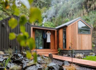 A modern tiny house with wood and black siding sits among rocks and greenery. Smoke rises from a chimney, and clothing hangs on a hook near the glass front door. A wooden walkway leads to the entrance.
