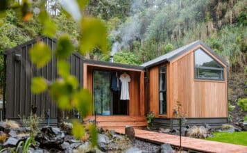 A modern tiny house with wood and black siding sits among rocks and greenery. Smoke rises from a chimney, and clothing hangs on a hook near the glass front door. A wooden walkway leads to the entrance.