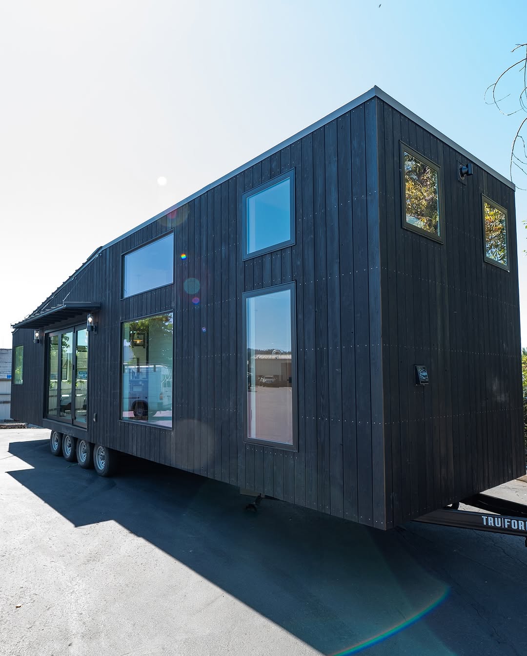 A sleek black tiny house on wheels with large windows, parked on a paved surface under a clear sky.