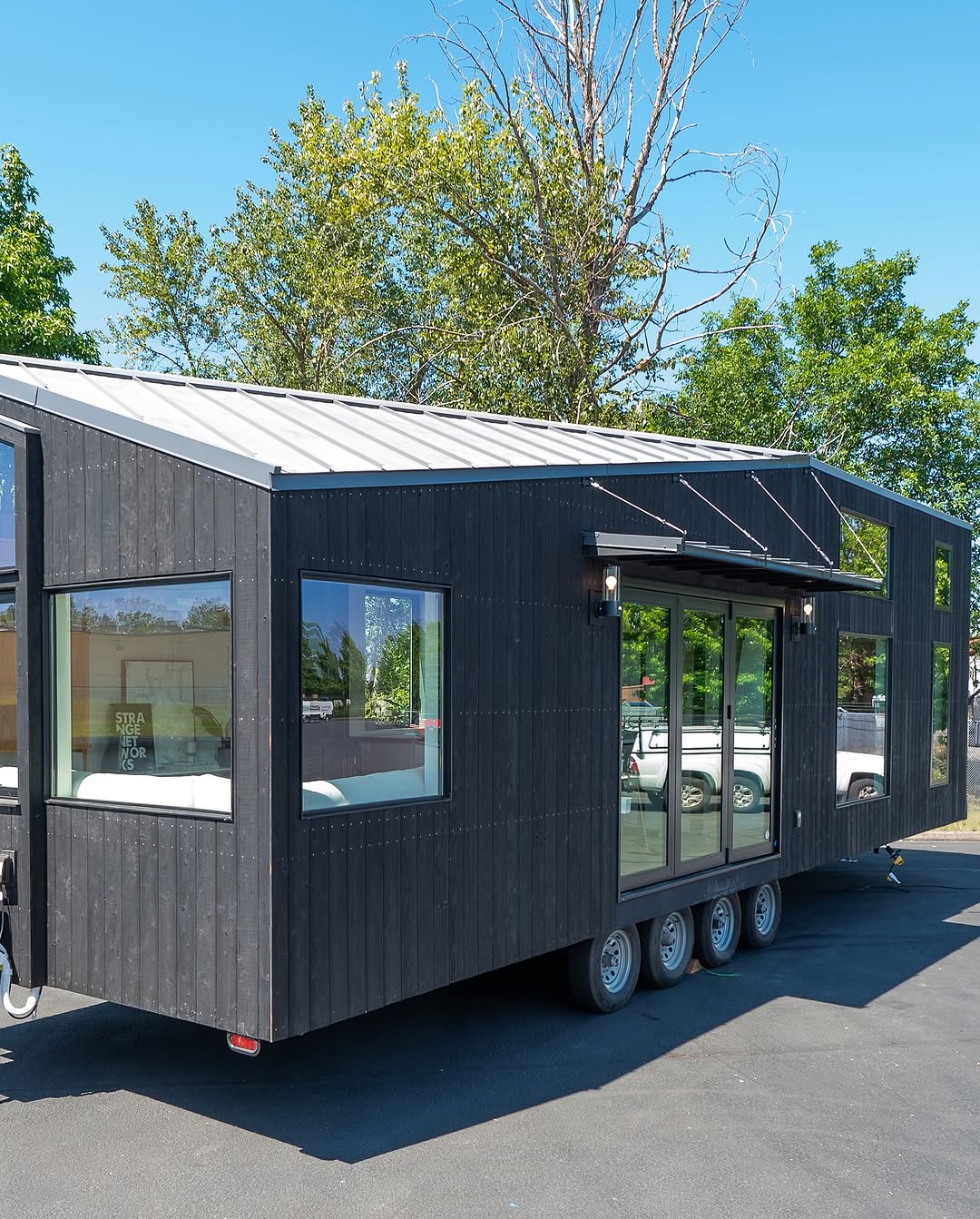 A modern black tiny house with large windows and sliding glass doors sits on a triple-axle trailer, parked on pavement. This stylish tiny house is surrounded by green trees beneath a clear blue sky.