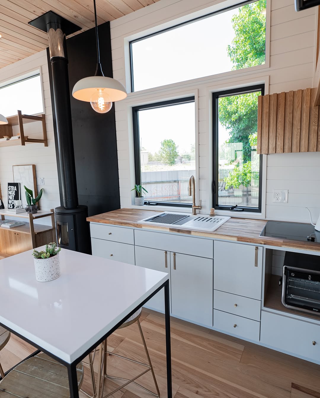 Modern tiny house kitchen with white cabinets, wood countertops, large windows flooding the space with natural light, and a small potted plant on a white island. A hanging light fixture and wood accents add warmth to this bright retreat.