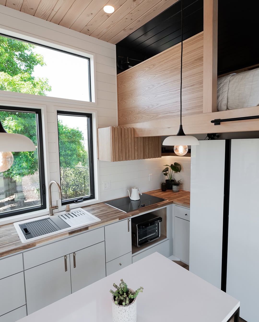 A modern, compact kitchen in a tiny house features light wood cabinets, white countertops, a sink by a large window, pendant lights, built-in appliances, a potted plant on the island, and a cozy loft bed above.