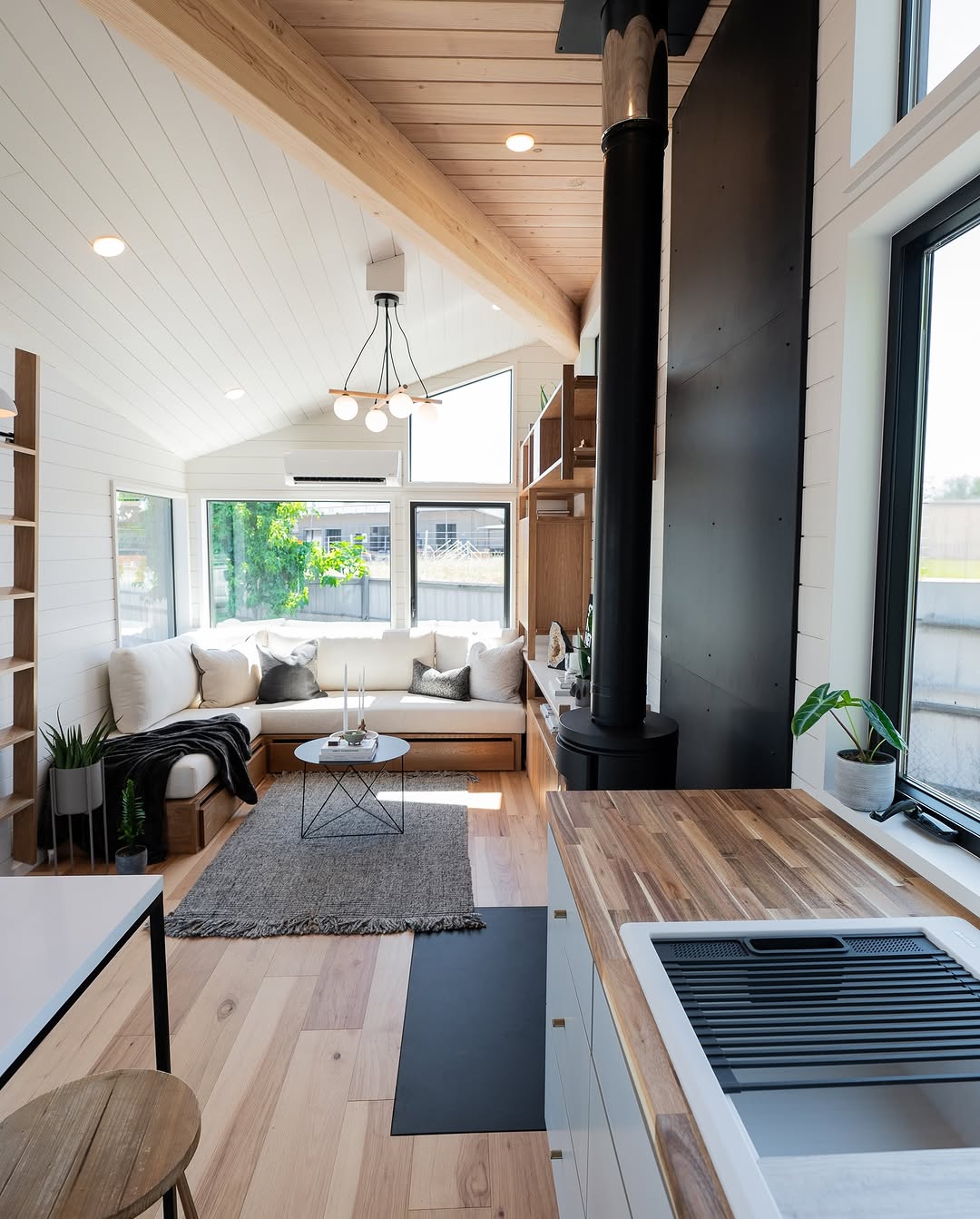A bright, modern tiny house interior features wood floors, a cozy corner sofa, coffee table, rug, large windows, a small wood stove, open shelving with houseplants, and a wooden countertop with sink in the foreground.
