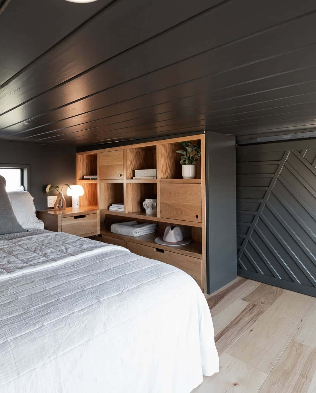 A cozy tiny house bedroom with a wooden bed, white bedding, light wood floors, and built-in shelves for books, decor, and towels. The dark walls and chevron-patterned sliding door add style to the compact space.