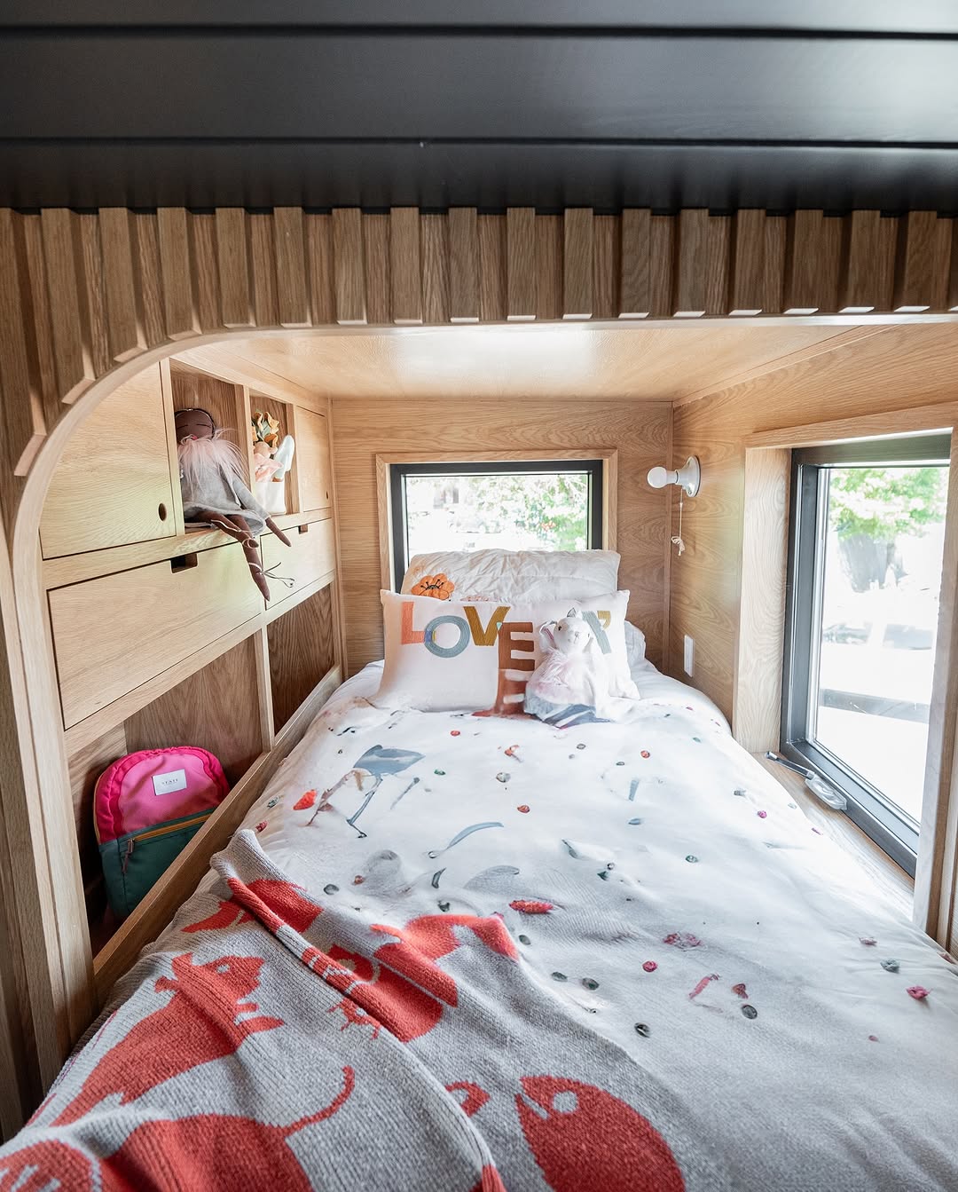 A cozy wooden nook bed reminiscent of a tiny house, with windows on both sides, animal-themed bedding, a LOVE pillow, a pink backpack in a cubby, and stuffed animals arranged on shelves above the bed.