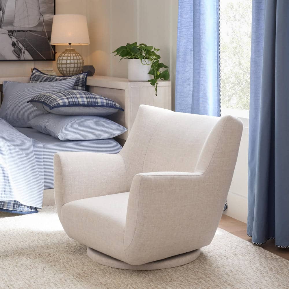 A cozy bedroom features a beige swivel armchair on a light rug, a neatly made bed with blue plaid and striped bedding, a bedside table with a lamp, and a potted plant near a window with blue curtains.