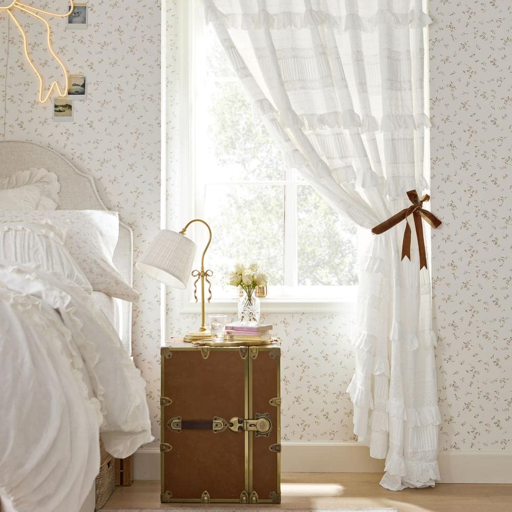 A cozy bedroom corner with floral wallpaper, a ruffled white curtain tied with a brown ribbon, and a vintage trunk used as a nightstand, topped with a lamp, books, and flowers beside a white bed.