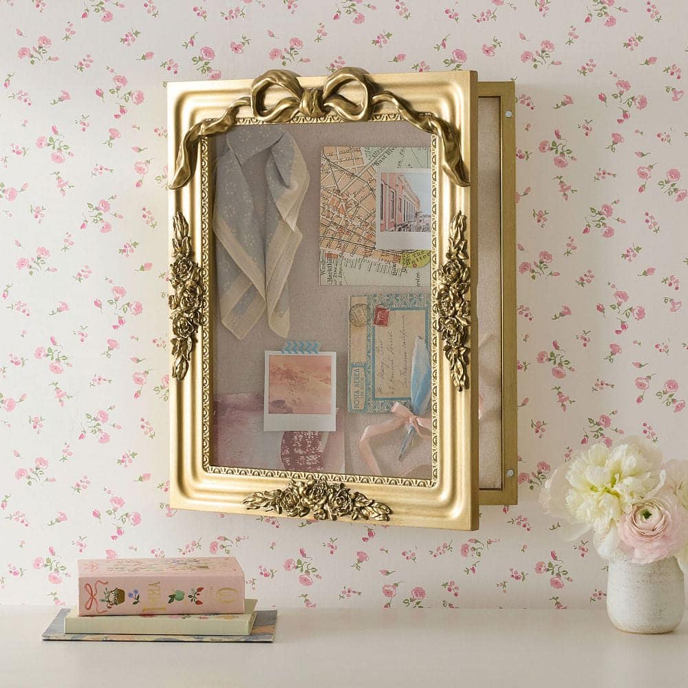 A gold ornate, framed shadow box with a bow at the top hangs on a floral wallpapered wall; inside are vintage maps, notes, ribbon, and fabrics. A vase of white flowers and a decorative book sit below.