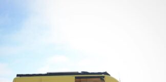 A yellow and black camper van is parked on sandy ground with its side door open, revealing a cozy, modern interior. The stylish camper van features white walls, wooden accents, and overlooks an open, empty outdoor area.