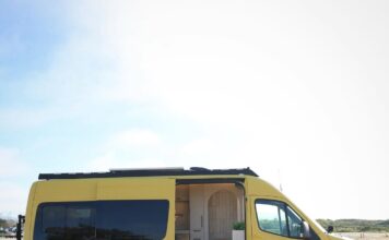 A yellow and black camper van is parked on sandy ground with its side door open, revealing a cozy, modern interior. The stylish camper van features white walls, wooden accents, and overlooks an open, empty outdoor area.