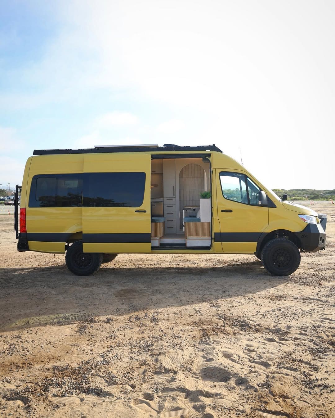 A yellow and black camper van is parked on sandy ground with its side door open, revealing a cozy, modern interior. The stylish camper van features white walls, wooden accents, and overlooks an open, empty outdoor area.