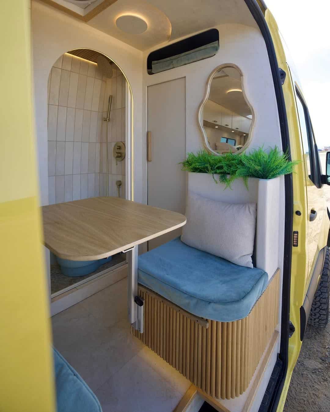 The cozy interior of a camper van features a small dining area with a wooden table, cushioned blue seats, a potted plant, an arched mirror, and a tiled shower visible in the background.
