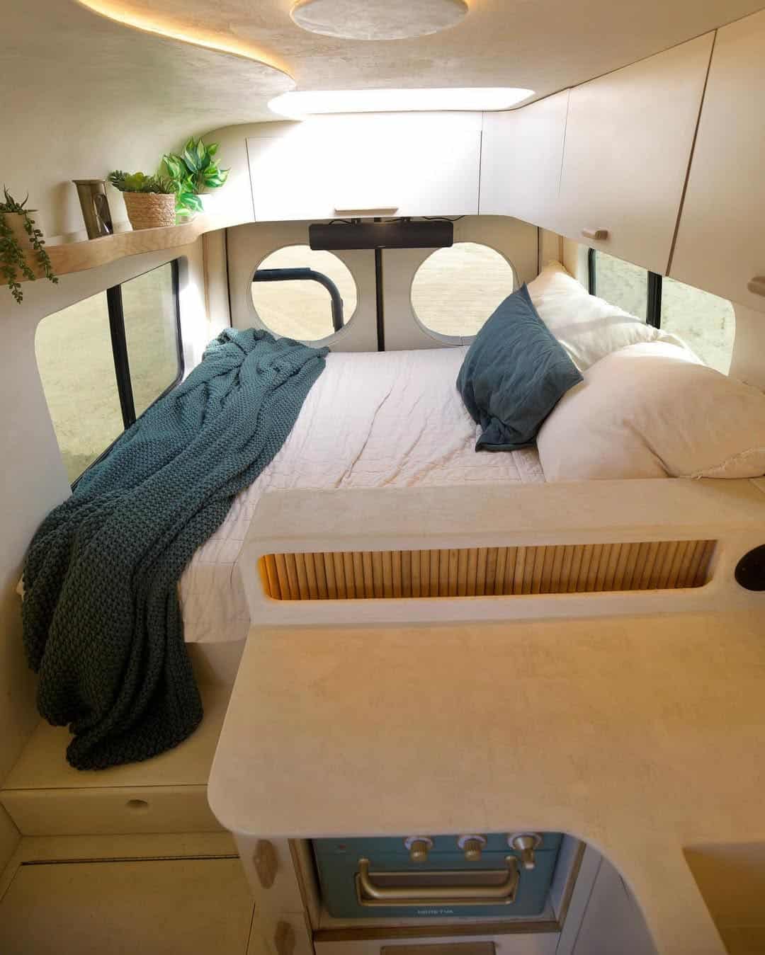 Cozy bed with pillows and a blue blanket inside a bright, modern camper van. Round windows, plants, and minimalistic decor create a comfortable, inviting camper van space.
