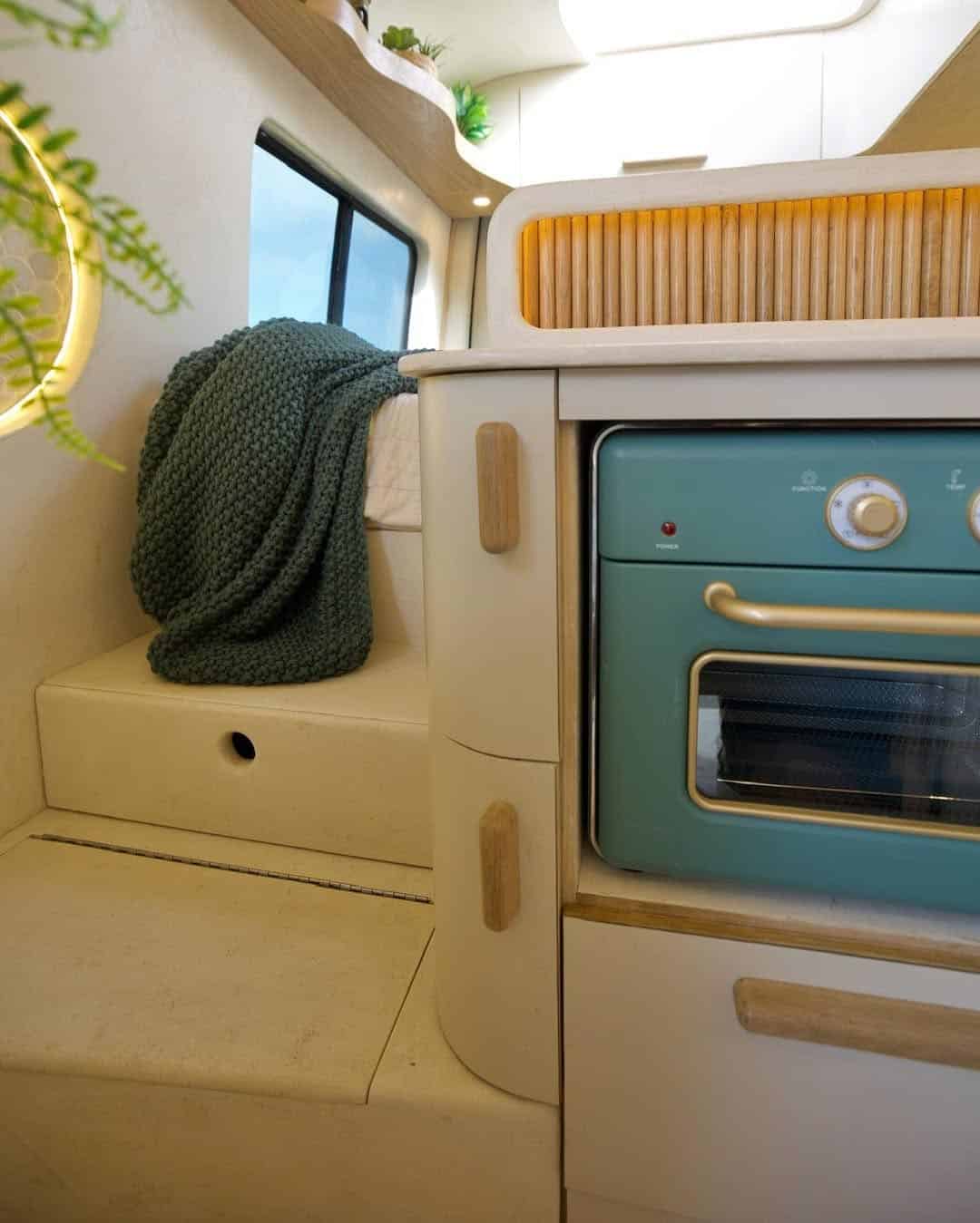 A cozy camper van interior featuring a teal oven, beige cabinetry, a green knitted blanket on a bench, a window, and plants decorating the corner.