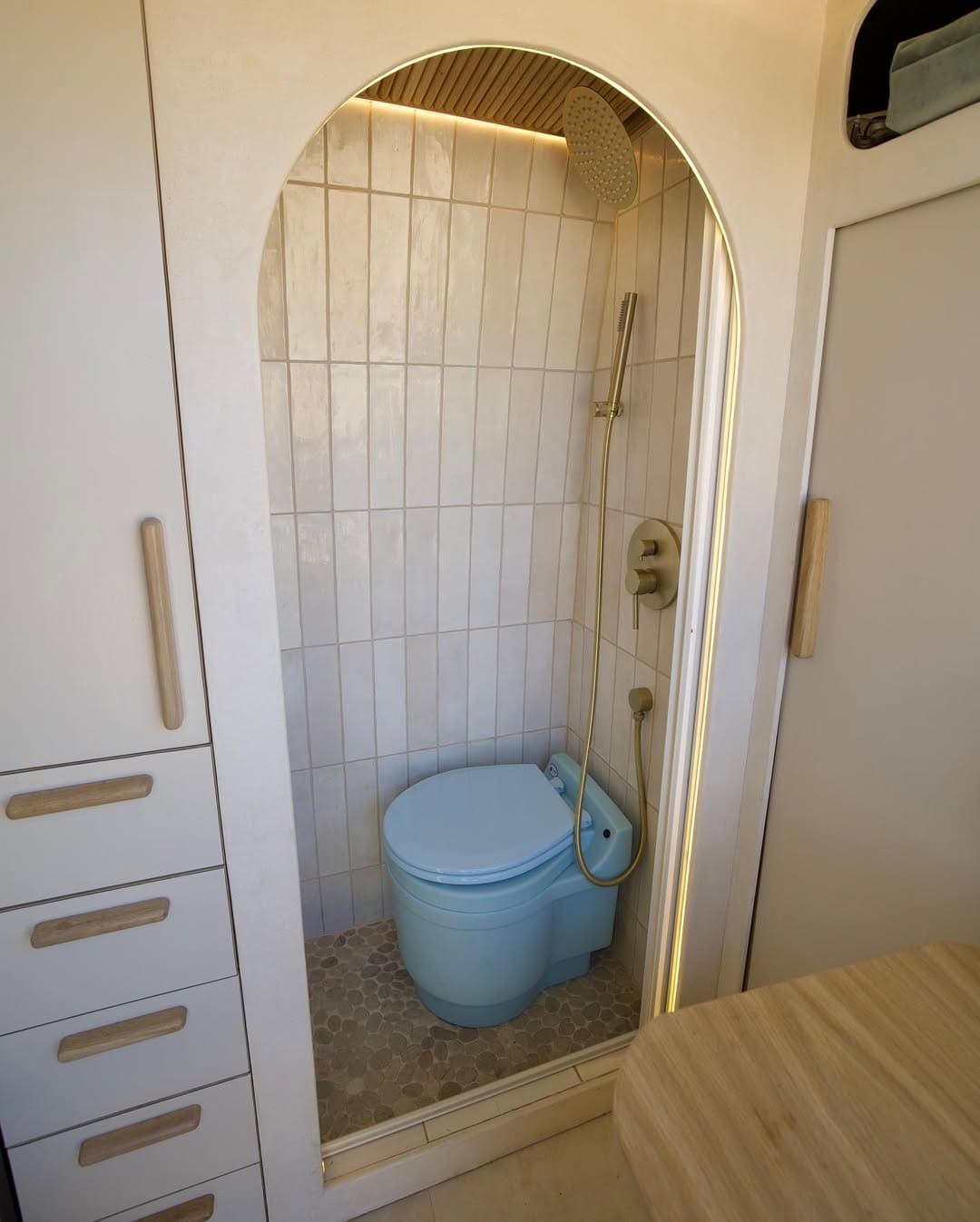 A compact camper van bathroom with a blue toilet and a showerhead inside a small, tiled shower area. Light wood cabinets and drawers are on the left, and a wooden countertop is partially visible on the right.
