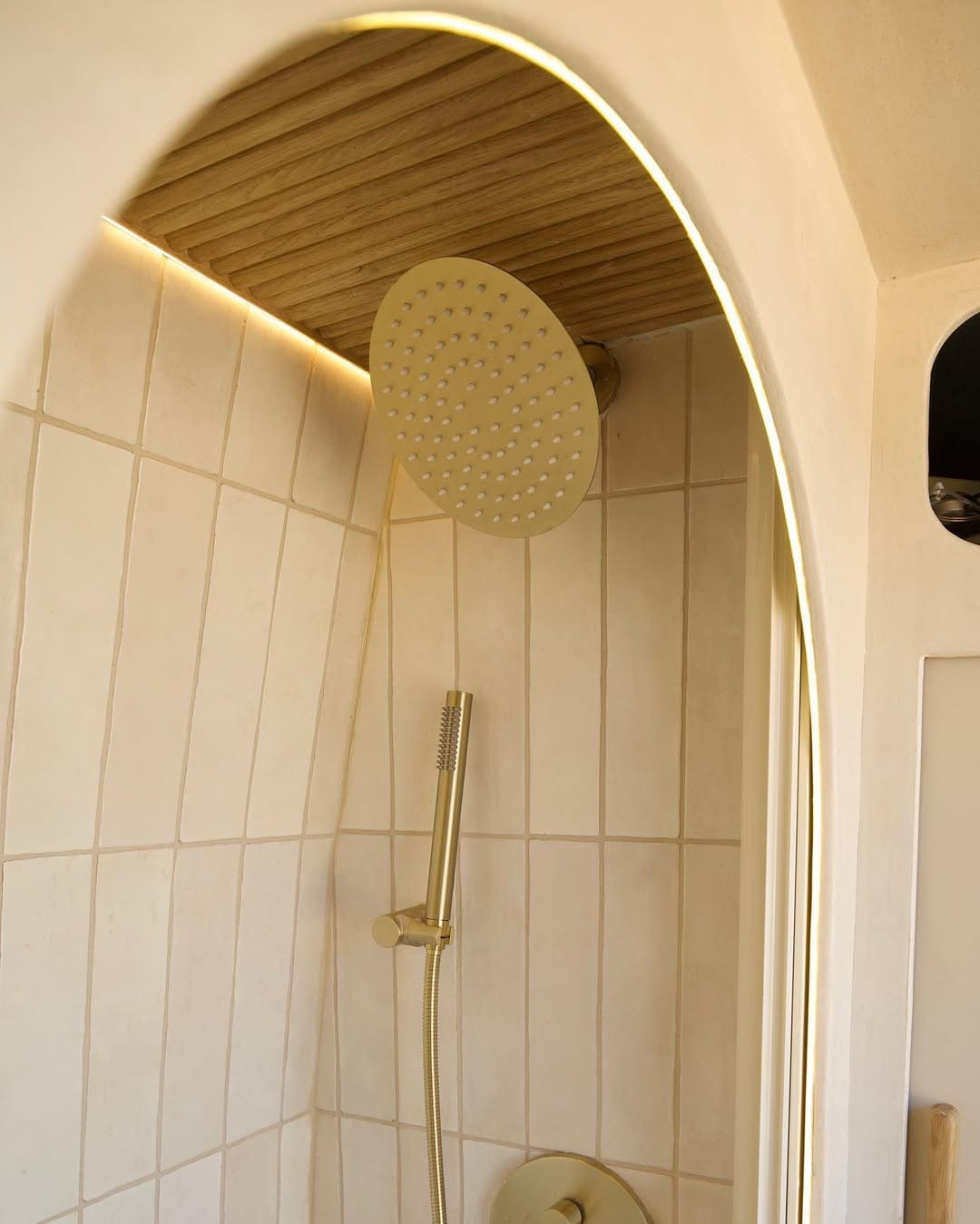 A modern shower in a camper van with a round, gold-colored rainfall showerhead, matching handheld sprayer, and light beige tiled walls. Soft lighting highlights wood paneling on the ceiling.
