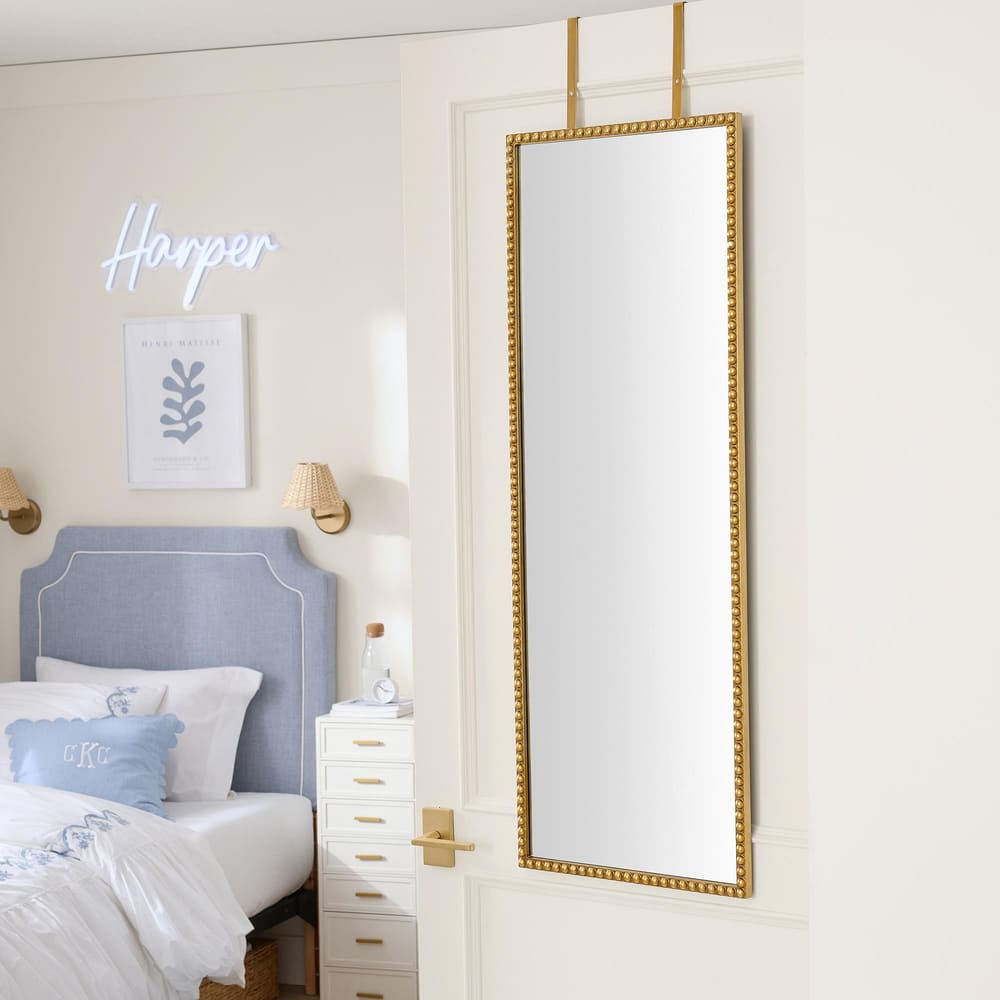 A gold-framed full-length mirror hangs on a white door in a bedroom with a blue bed, white bedding, a small white dresser, and a neon sign reading Harper on the wall.