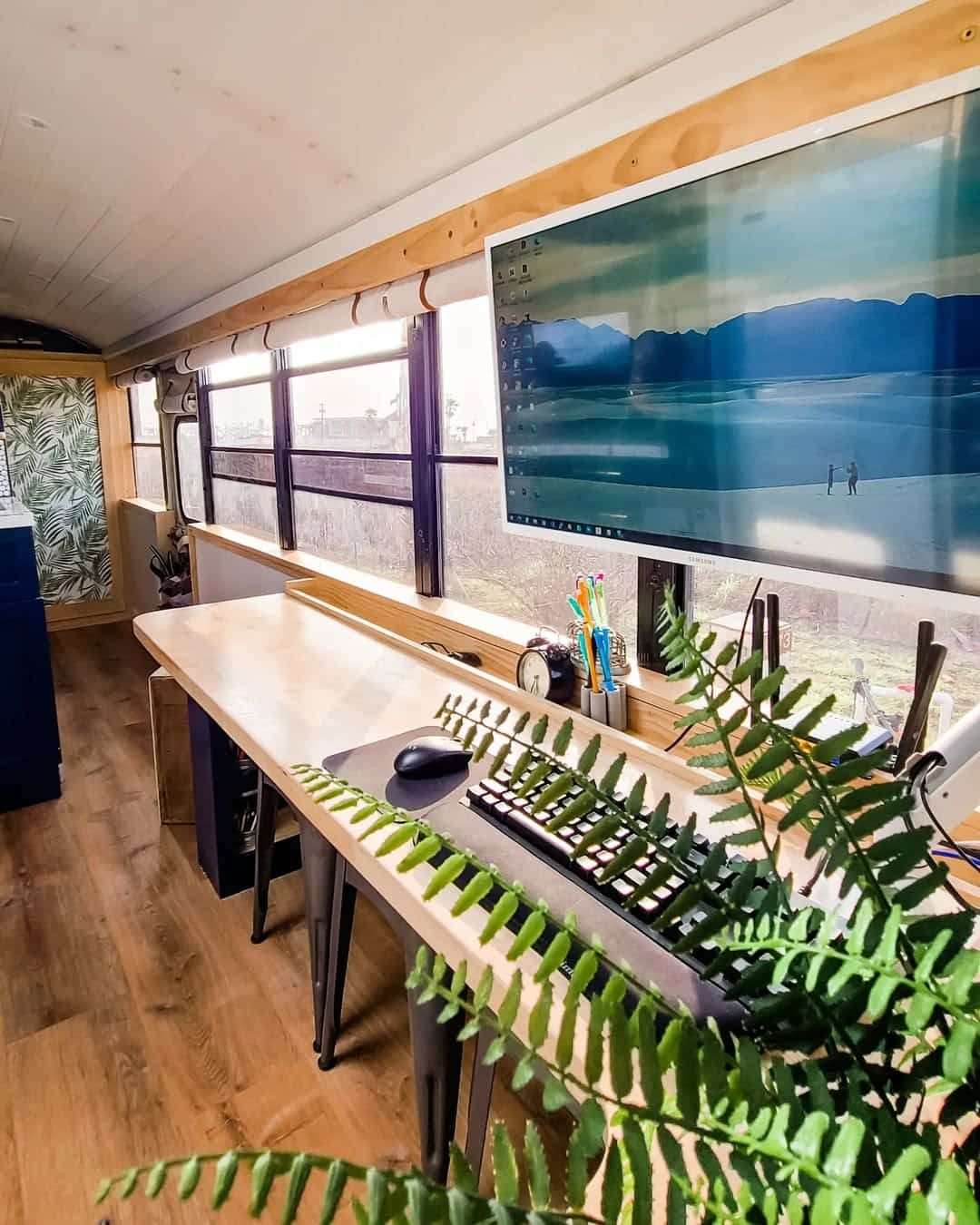 A modern skoolie workspace inside a converted bus features a wooden desk, large wall-mounted monitor, keyboard, mouse, office supplies, and a green fern in the foreground, with large windows offering a scenic outdoor view.