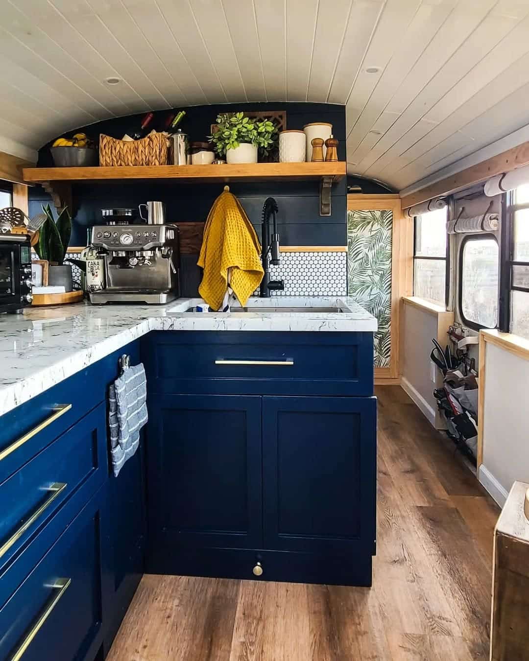 A cozy, modern skoolie kitchen inside a renovated bus features navy cabinets, a marble countertop, a black faucet, open wooden shelves with plants and decor, and wood floors. Sunlight streams in through large bus windows.
