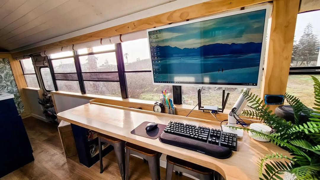 A workspace inside a converted skoolie bus features a large monitor above a long wooden desk with keyboard, mouse, and stationery. Expansive windows line the wall, filling the space with natural light and views of trees and grass outside.