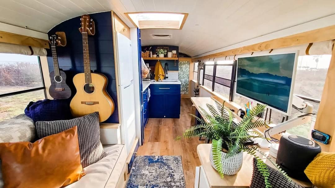 Interior of a cozy skoolie with wooden floors, blue cabinets, a kitchen area, hanging guitars, a sofa with cushions, large windows, a TV, a potted plant, and various homey decorations.