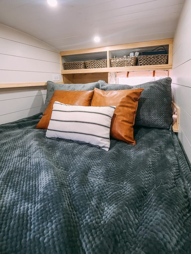 Cozy skoolie bed with a textured dark green blanket, two leather accent pillows, a striped pillow, and soft headboard pillows. Wooden shelves above hold baskets and decor, all softly lit by warm recessed lights.
