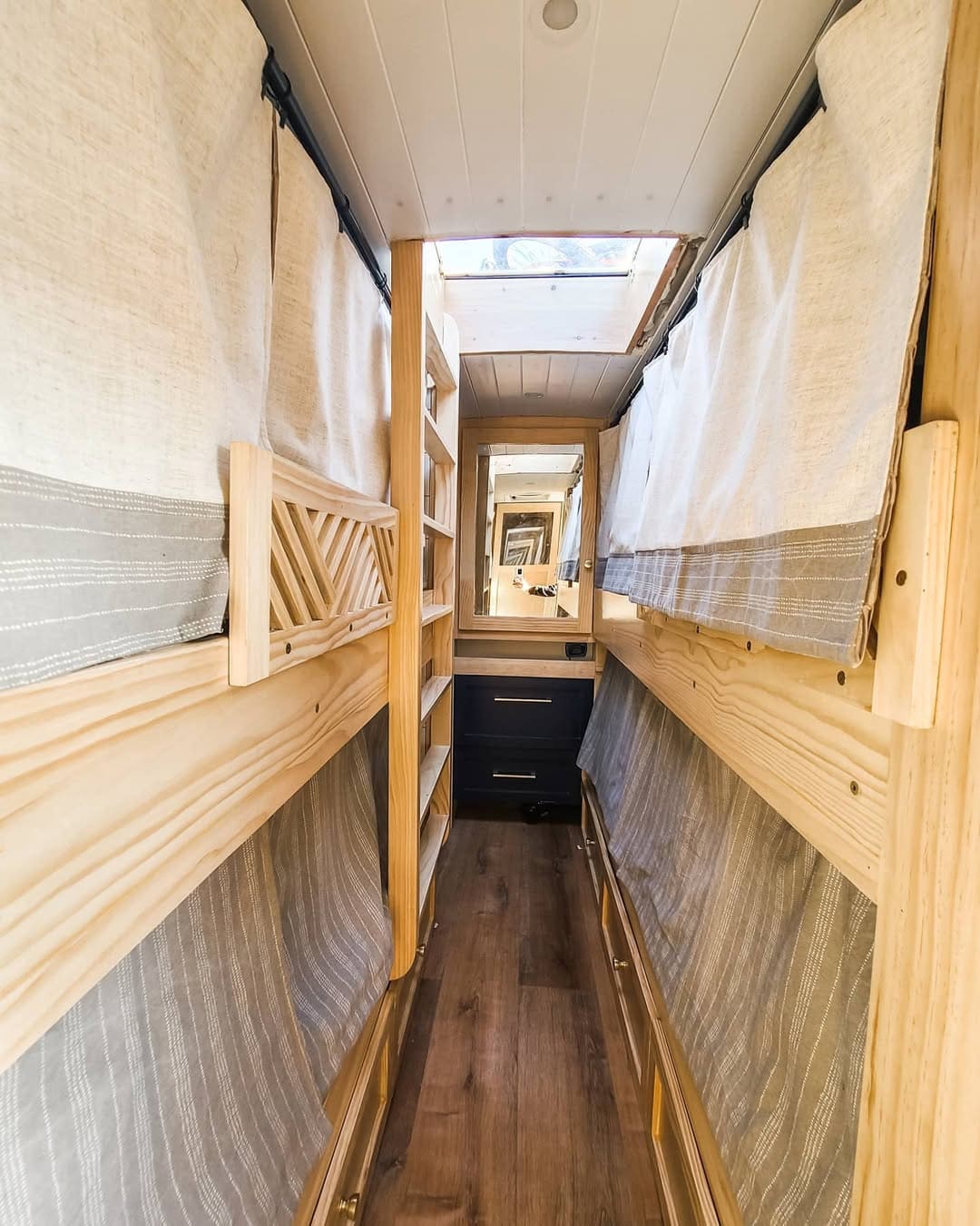 A narrow skoolie interior features wooden bunk beds on both sides, light curtains for privacy, shelves at the end, dark under-bed drawers, and a skylight letting in natural light above.