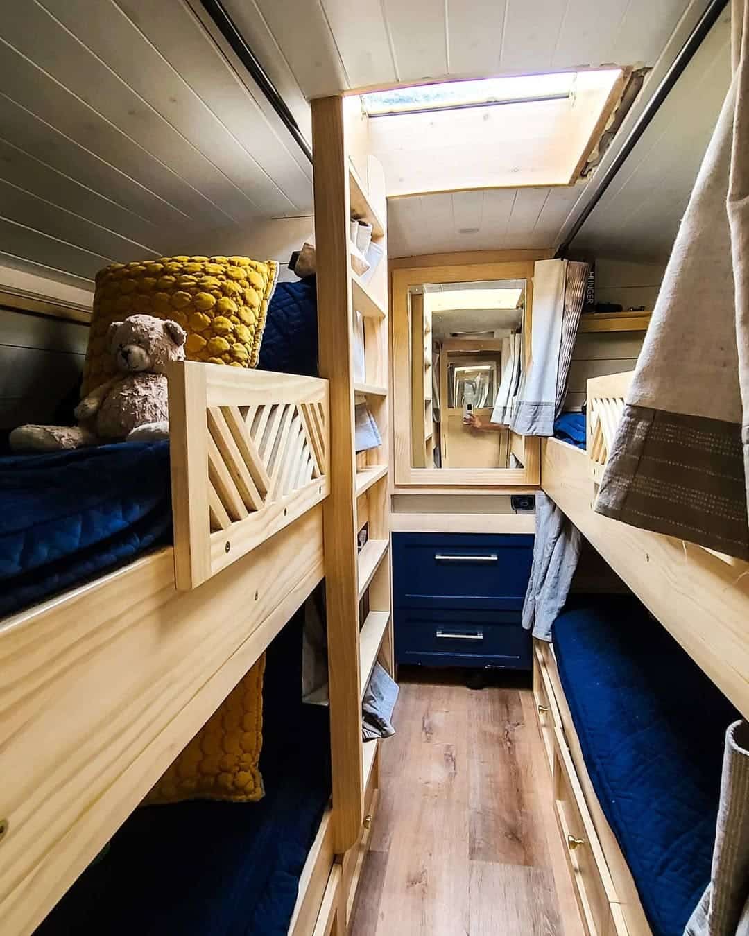 A cozy skoolie hallway with wooden bunk beds on both sides, blue bedding, a yellow cushion, a teddy bear, built-in drawers, and a mirror at the end reflecting the space. Natural light streams in from a skylight above.