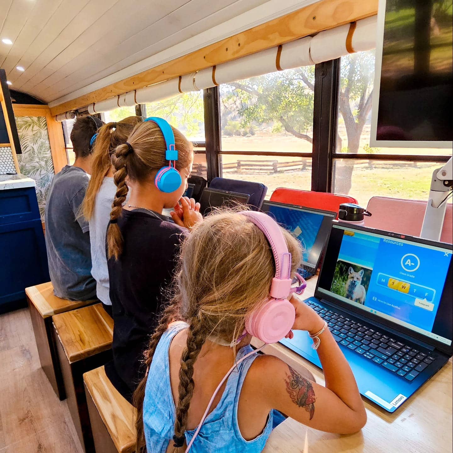 Three children wearing colorful headphones sit at wooden desks in a bright room with large windows, working on laptops. Trees and a fence outside suggest this could be a skoolie classroom or mobile learning environment.