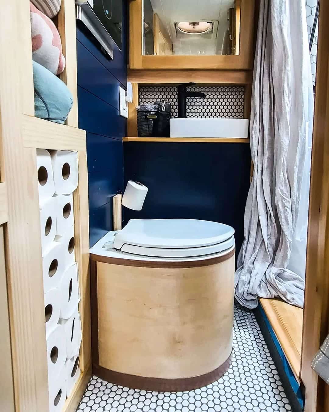Compact skoolie bathroom with a wooden composting toilet, stacked toilet paper rolls, small sink and mirror above, hexagon tile floor, and a striped curtain partially open to a window.