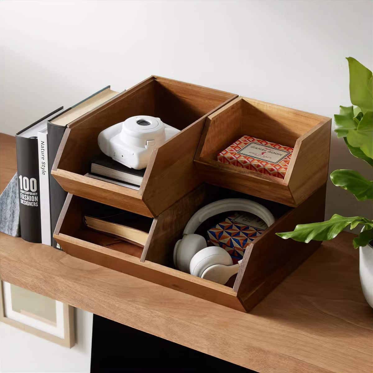 A wooden organizer with multiple compartments holds a camera, books, a box, headphones, and other small items—perfect for storage renters seeking tidy spaces. It sits on a wooden desk next to a plant and more books.